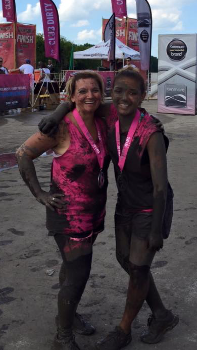 Two mud-covered women in pink shirts and medals pose after a muddy obstacle race.