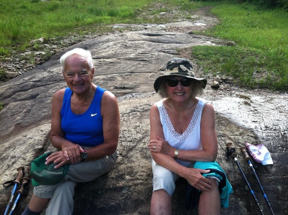 Two older adults smiling, resting on a rock path. Man in blue, woman in white tank top and hat. Hiking poles nearby.