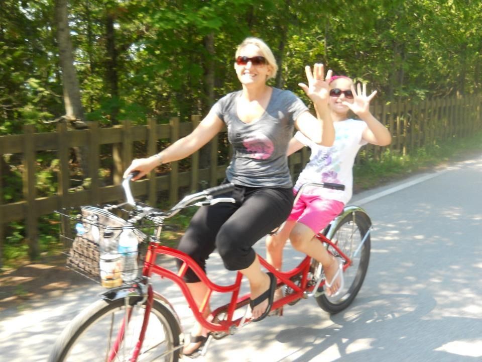 Woman and girl riding a red tandem bicycle on a path, waving, sunny day.