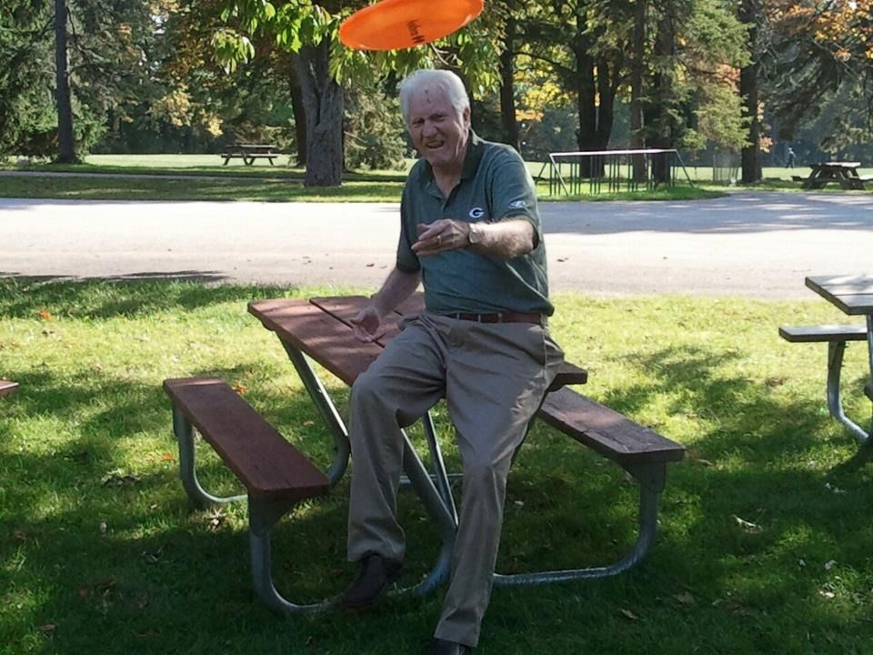 Older man in green shirt and tan pants playing frisbee at a park, smiling.
