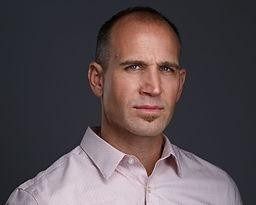 Man with short hair, wearing a pink collared shirt, looking directly at the viewer.