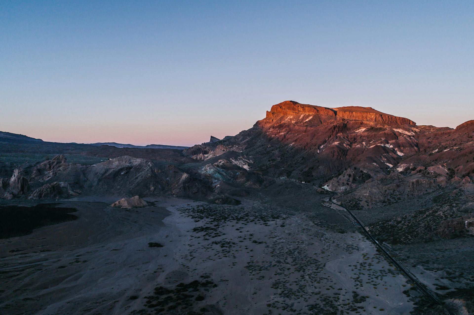 Mountain landscape at sunrise. Orange light on the peaks, road, and vast gray/brown terrain. Clear blue sky.