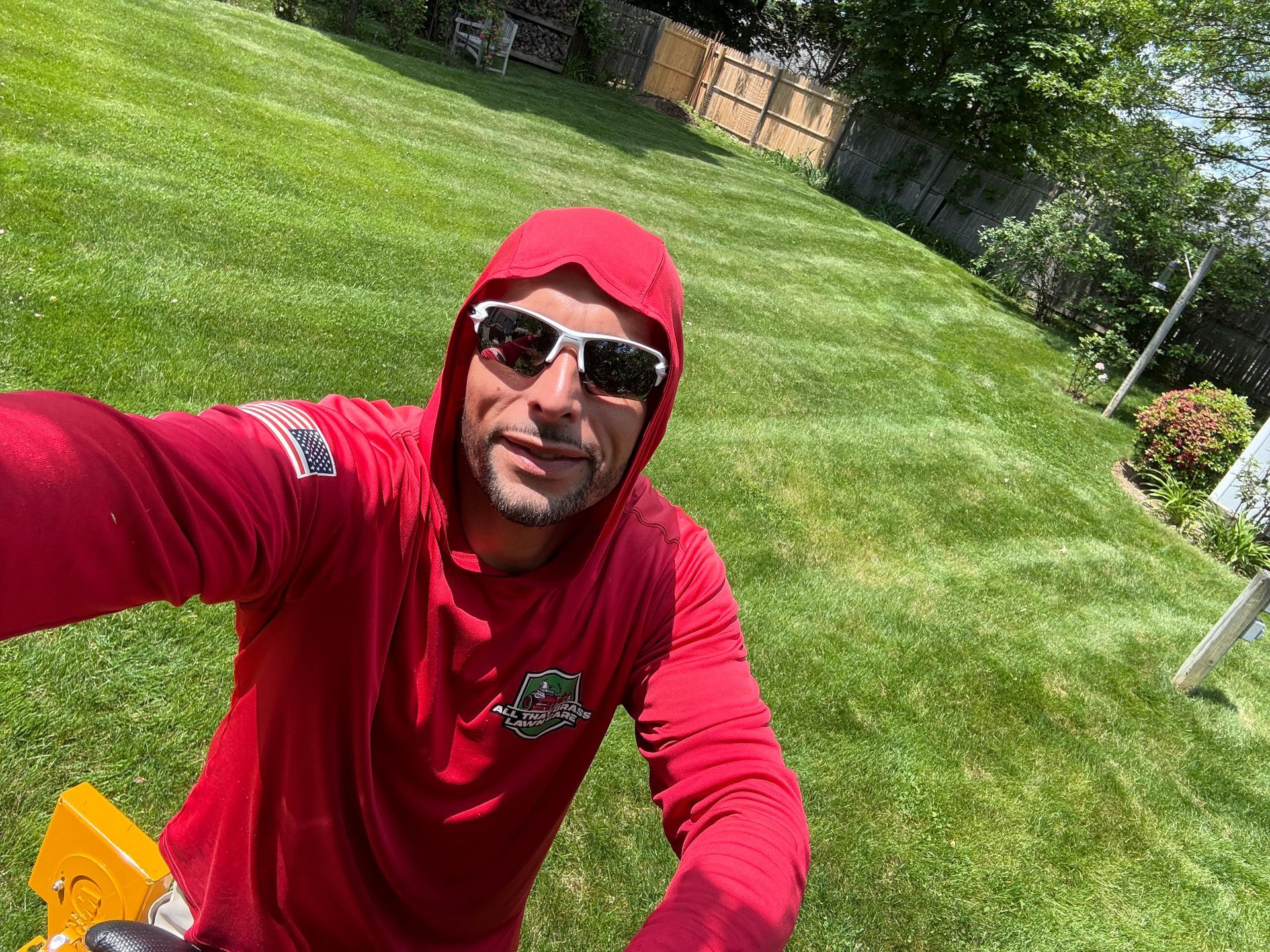 A man in a red hoodie and sunglasses is taking a selfie in a lush green yard.