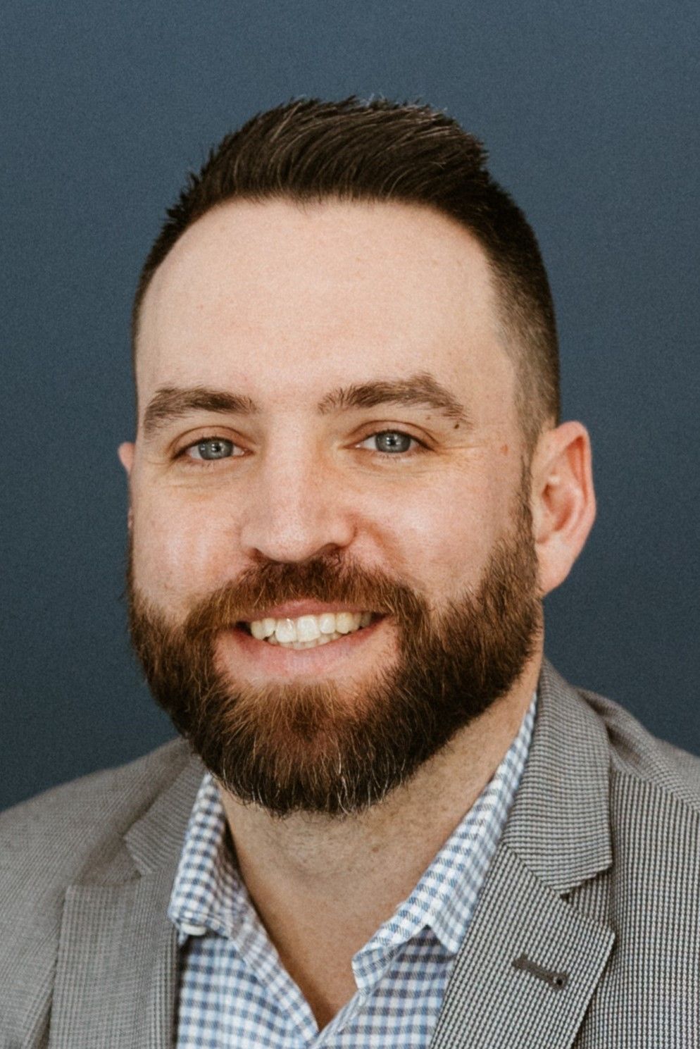 Man in light blue checkered shirt smiling, leaning against a building.