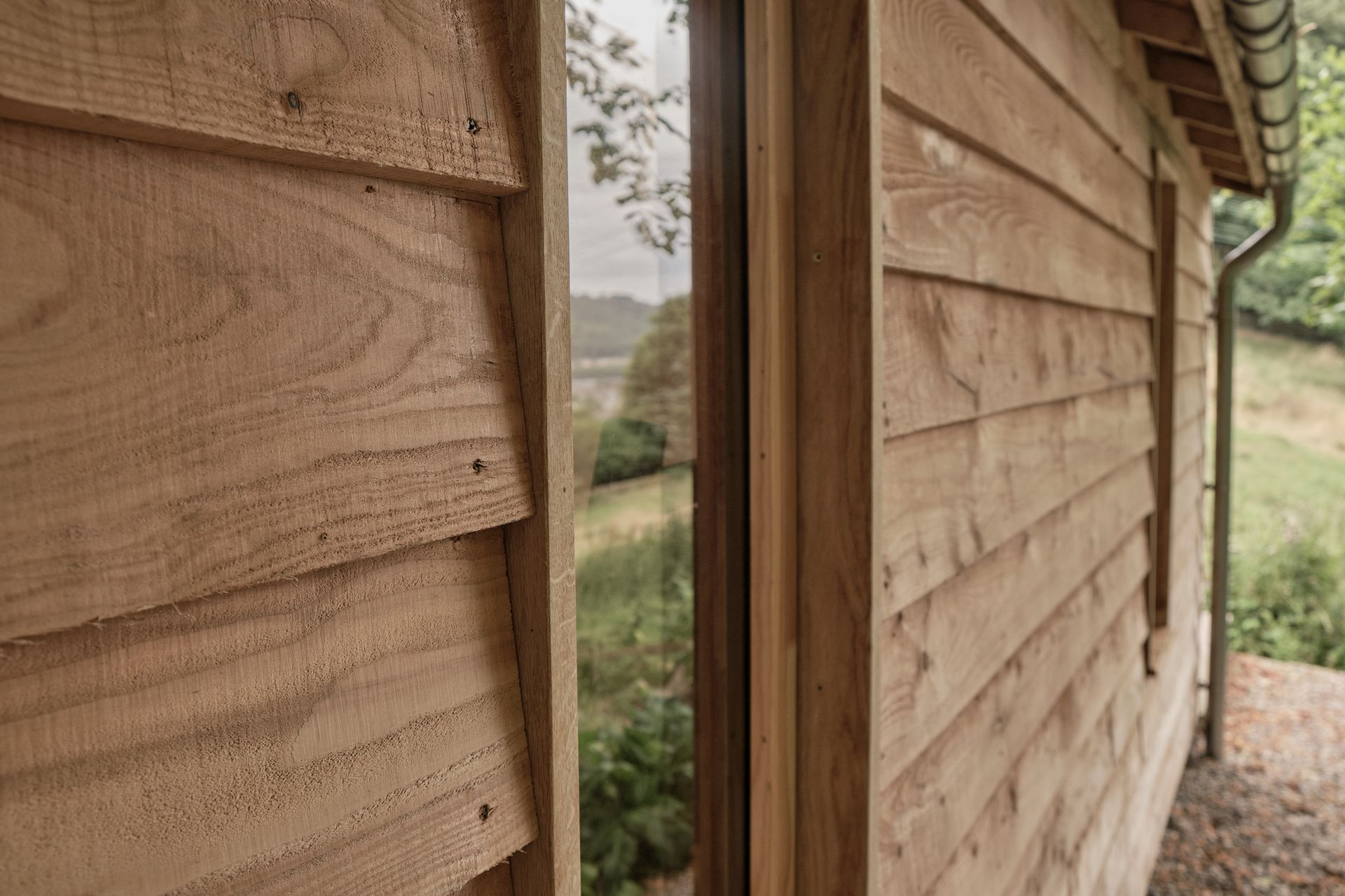 cabin wall with wooden window and rough sawn larch cladding. galvanised steel gutter and downpipe in distance