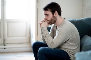 A Sad Man With A Beard Is Sitting On A Couch With His Hands Folded — Naturopath Australia In Mermaid Beach, QLD