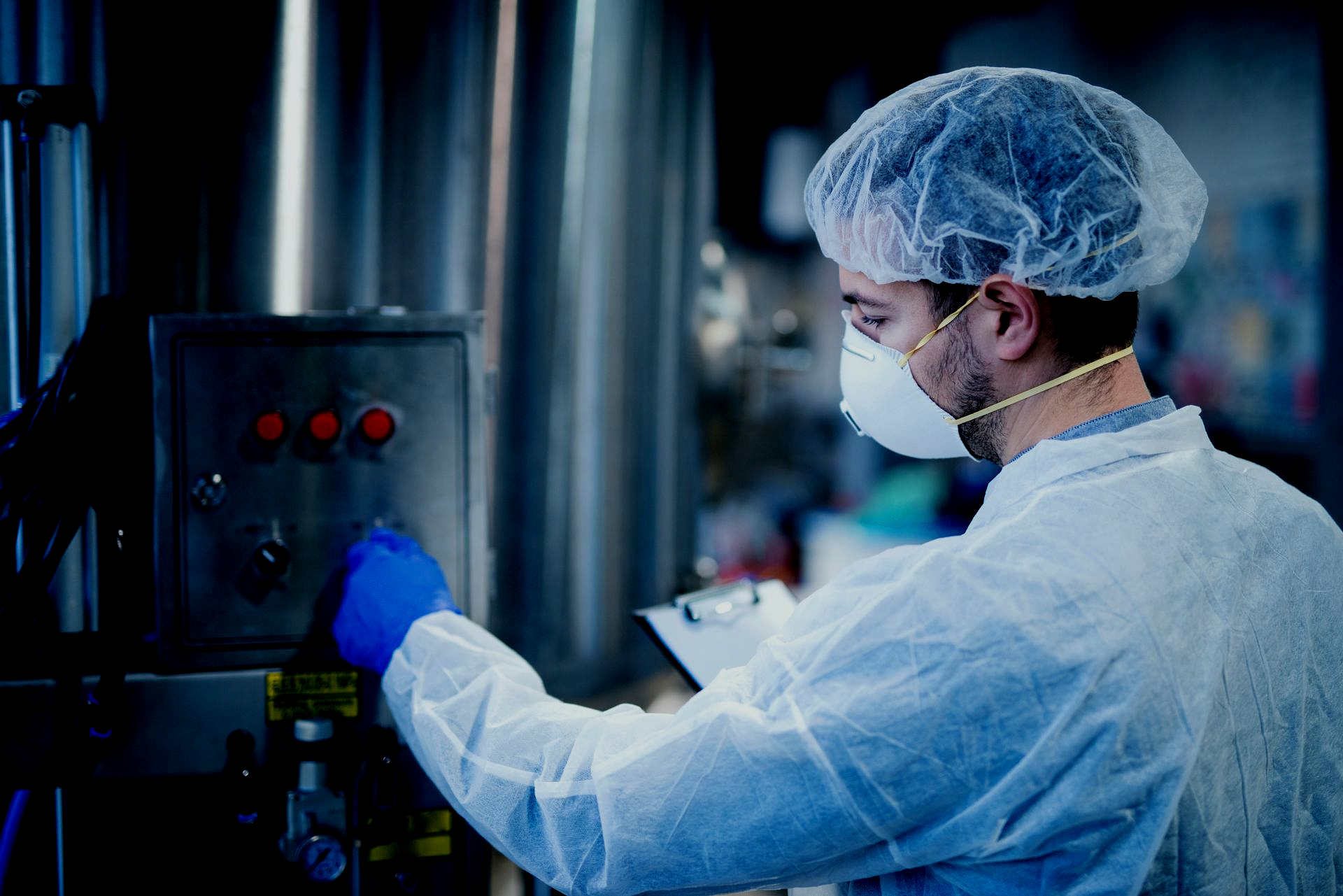 Person in protective gear operating machinery in a factory, holding a clipboard.