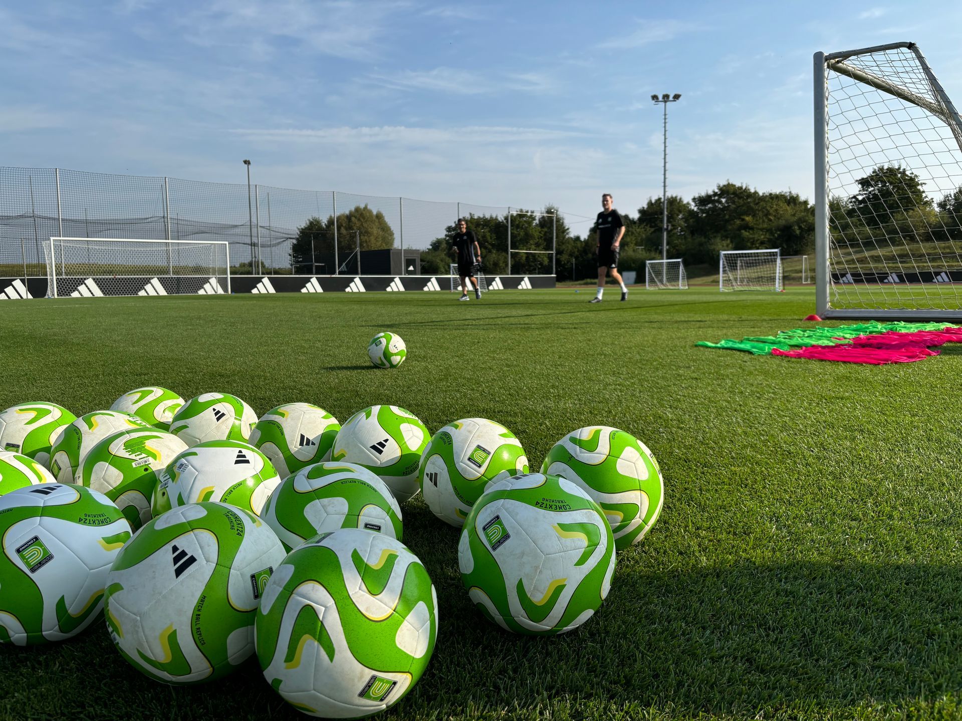 Soccer balls on a field near a goal, with a player and training equipment in the background