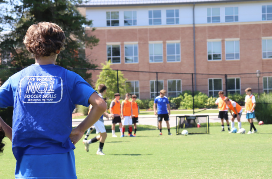 Coach in blue watches soccer practice on a sunny field outside a school building