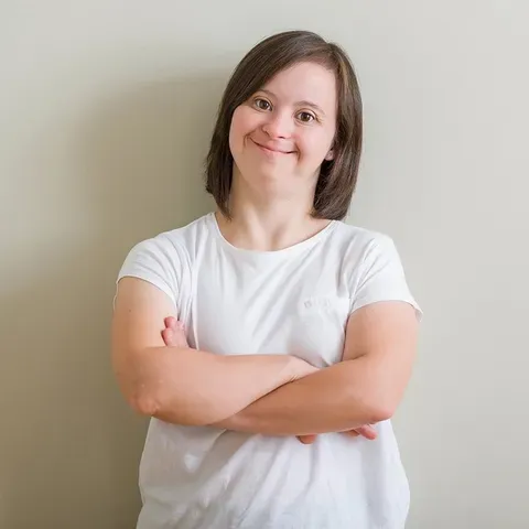 A woman with down syndrome is standing with her arms crossed and smiling.