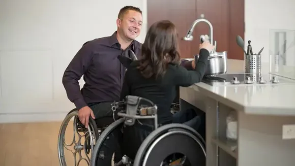 A man in a wheelchair is standing next to a woman in a wheelchair in a kitchen.