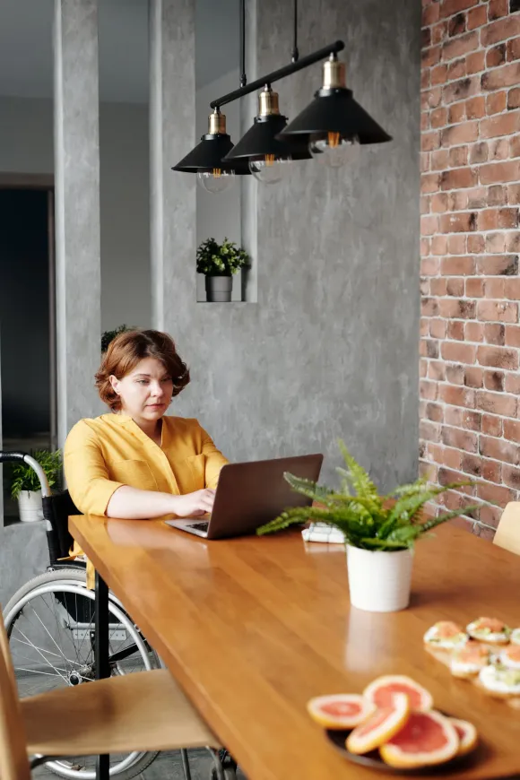 A woman in a wheelchair is sitting at a table using a laptop computer.