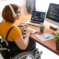 A woman in a wheelchair is sitting at a desk with a laptop and a computer.