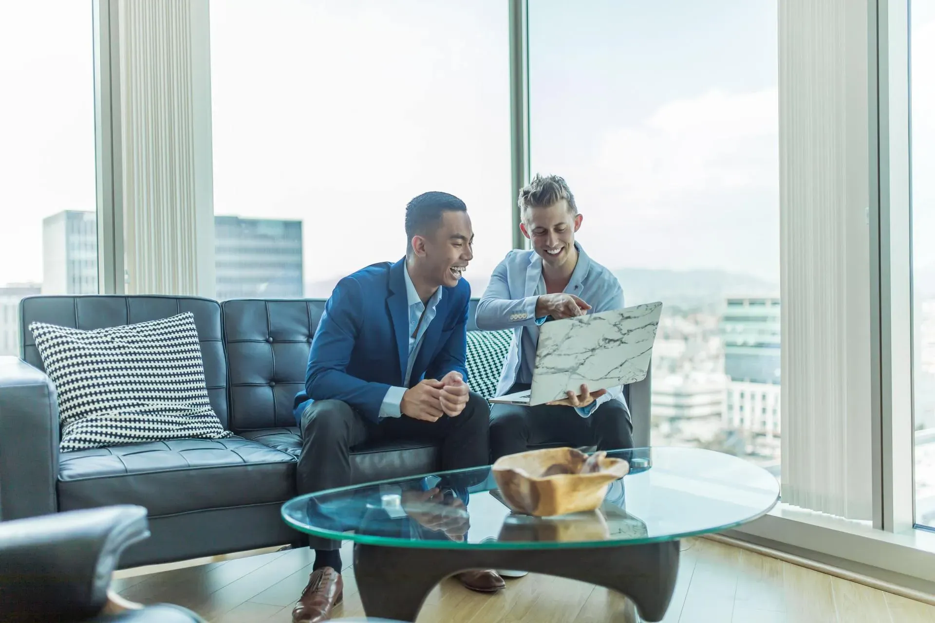 Two men are sitting on a couch in a living room looking at a laptop.