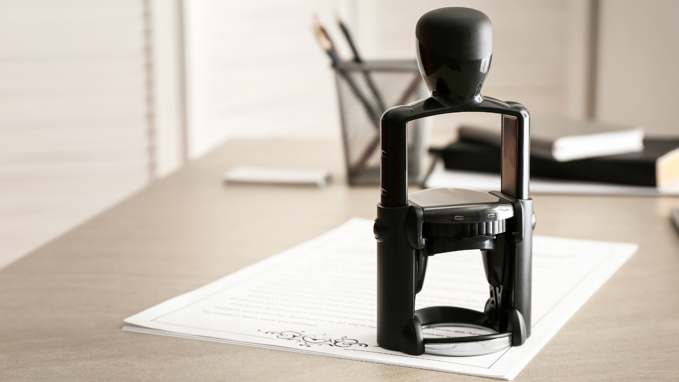 A black self-inking office stamp rests on a blank document on a desk next to a pen holder and notebooks.