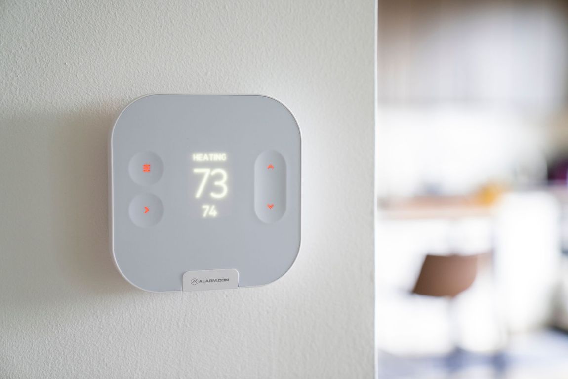 Wall-mounted gray thermostat displaying 73°, on a white wall with a blurred room in the background