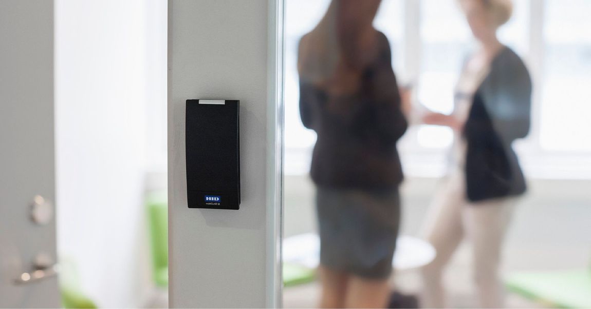 Black wall-mounted access device beside a blurred office doorway with two people in the background