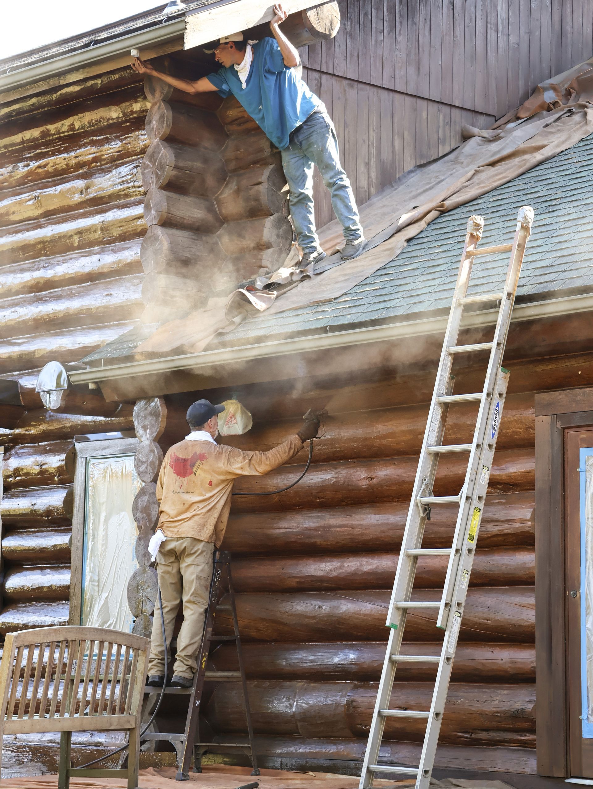 Two men are staining on a log cabin with a ladder.