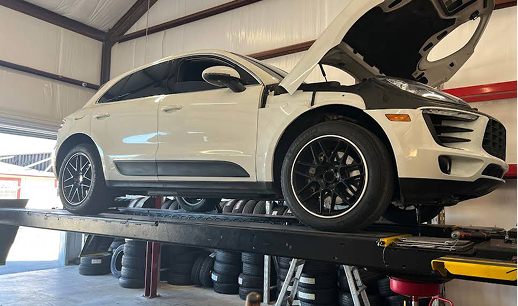 White Porsche SUV on a lift with the hood open in a mechanic's shop, black wheels.