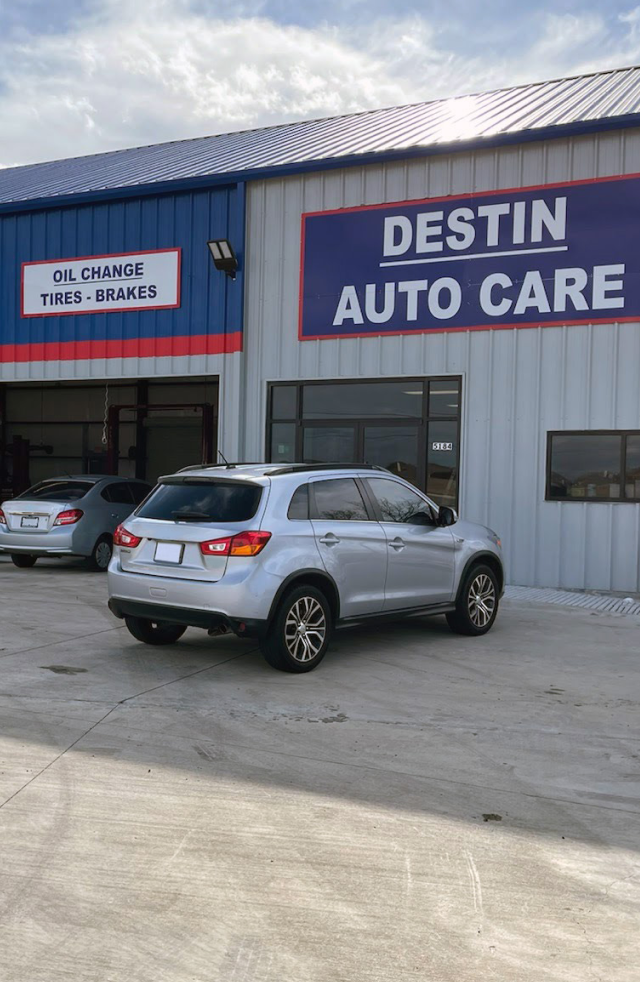 Silver SUV parked outside Destin Auto Care. Building has blue, red, and white trim. Gray sky.