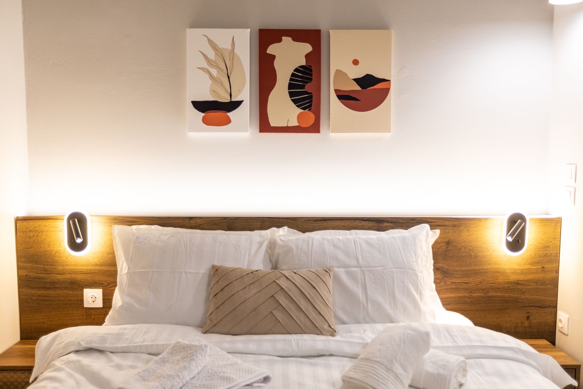 Bedroom with a bed, wooden headboard, artwork, sconces, and neutral colors.