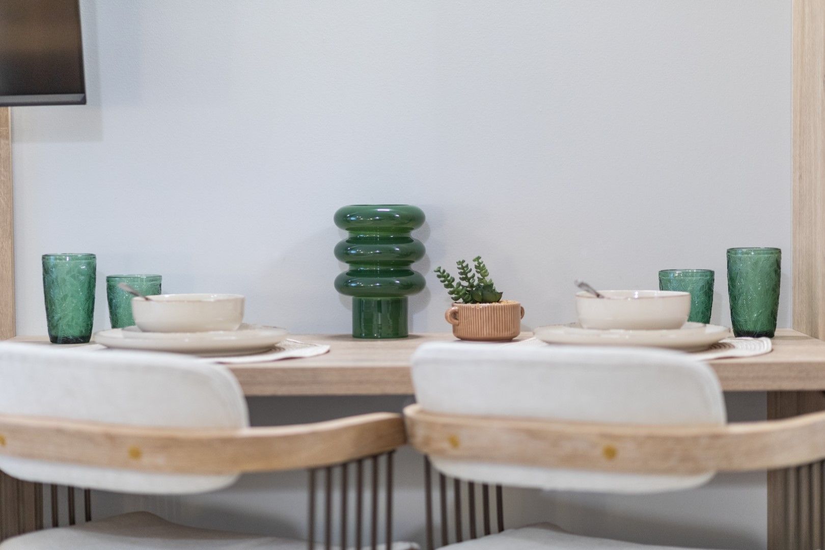 Dining table setting: white chairs, beige dishes, green glasses, and a decorative vase.