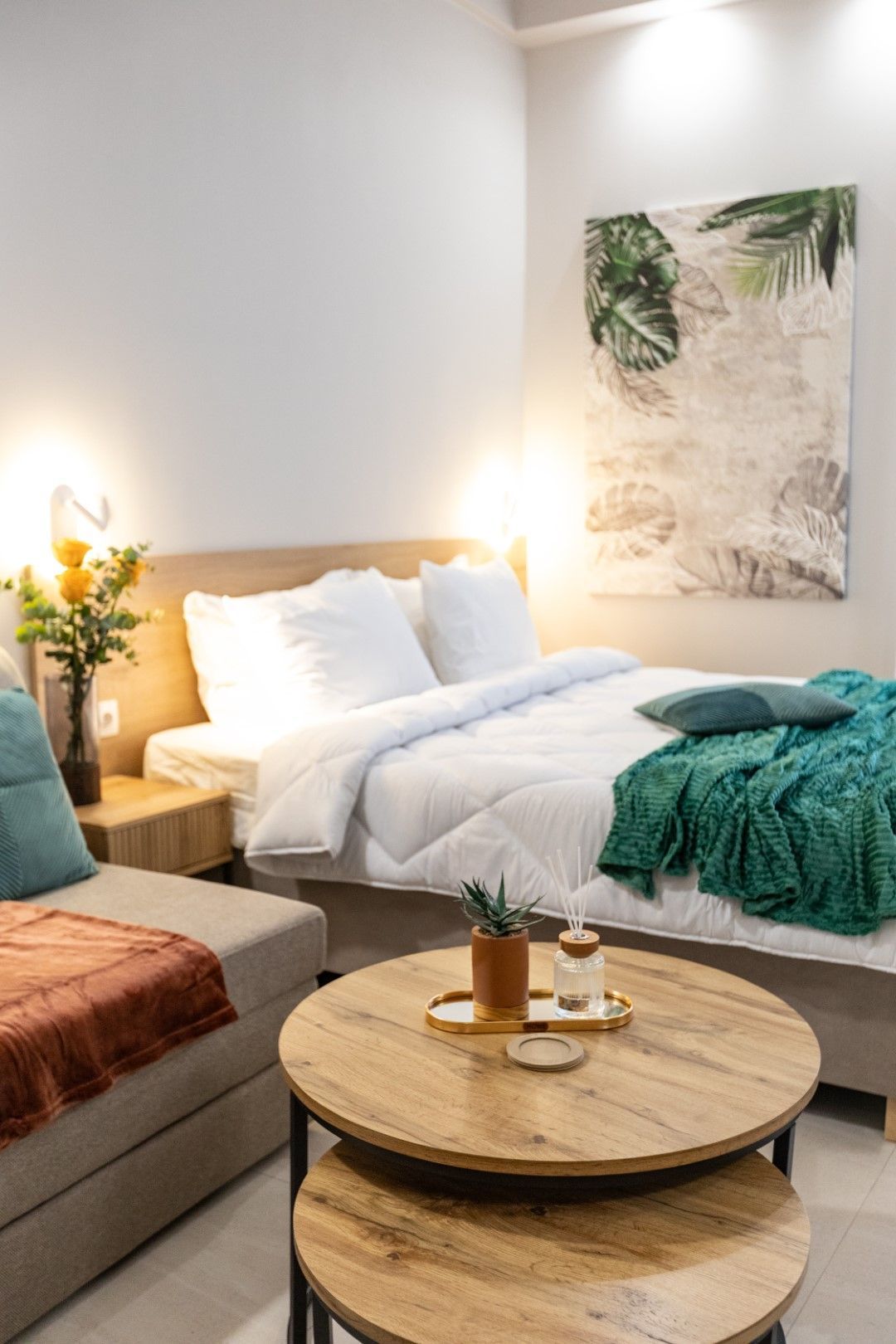 Cozy bedroom with a bed, small sofa, and wooden side table. Green blanket, pillows, and a plant add color.