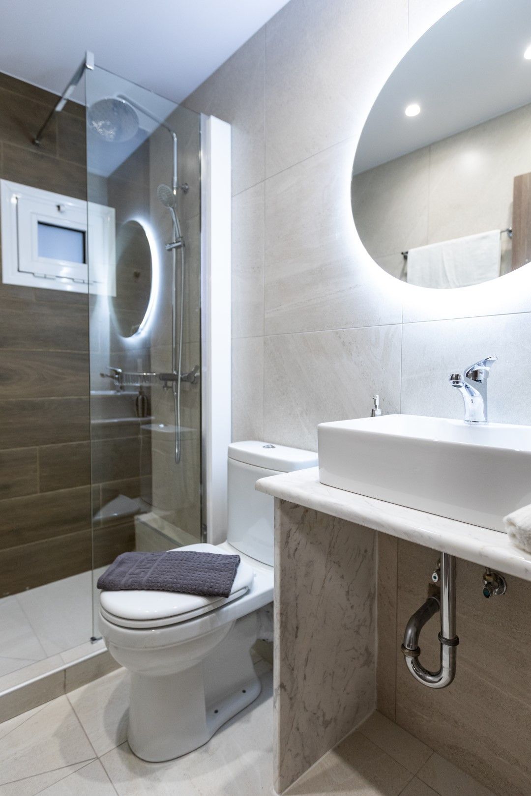 Modern bathroom with glass shower, toilet, and sink. White and beige tones, with a circular mirror.