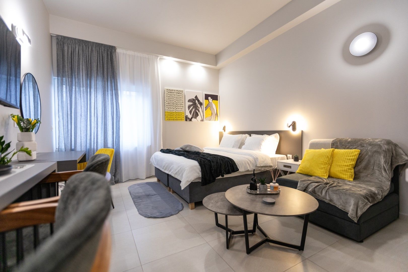 Modern hotel room with bed, sofa, desk, and round coffee tables. Gray, white, and yellow decor.