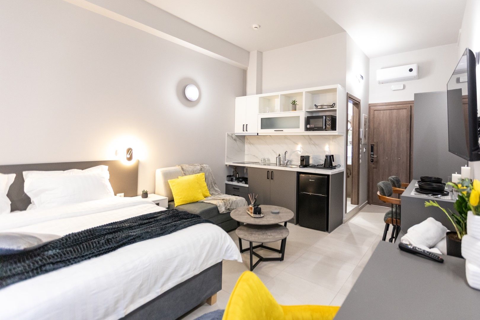 Modern studio apartment with bed, small kitchen, and seating area. Gray and white color scheme, yellow accents.