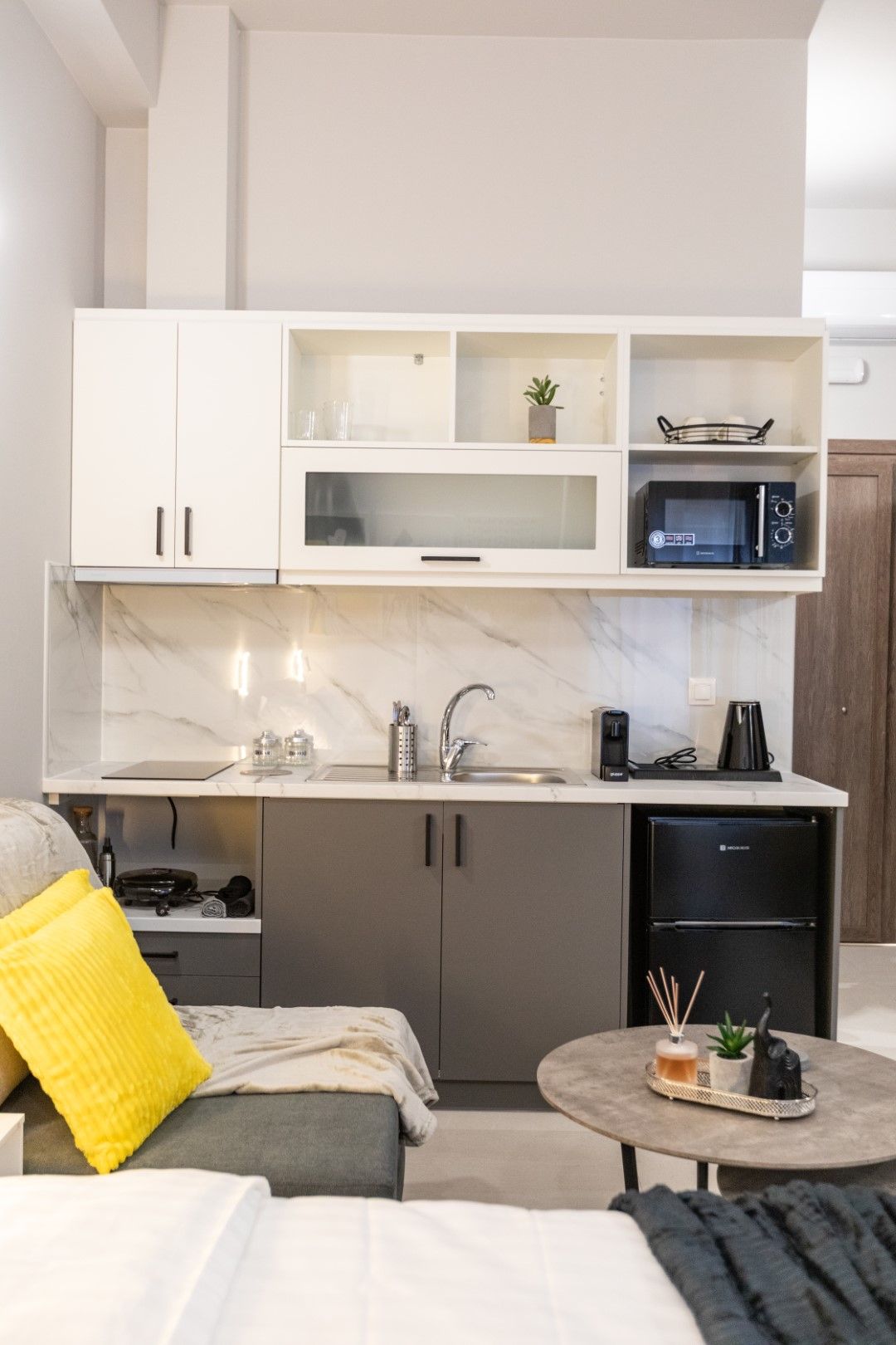 Small kitchen area with cabinets, sink, microwave, and mini-fridge. Grey, white, and wood tones.