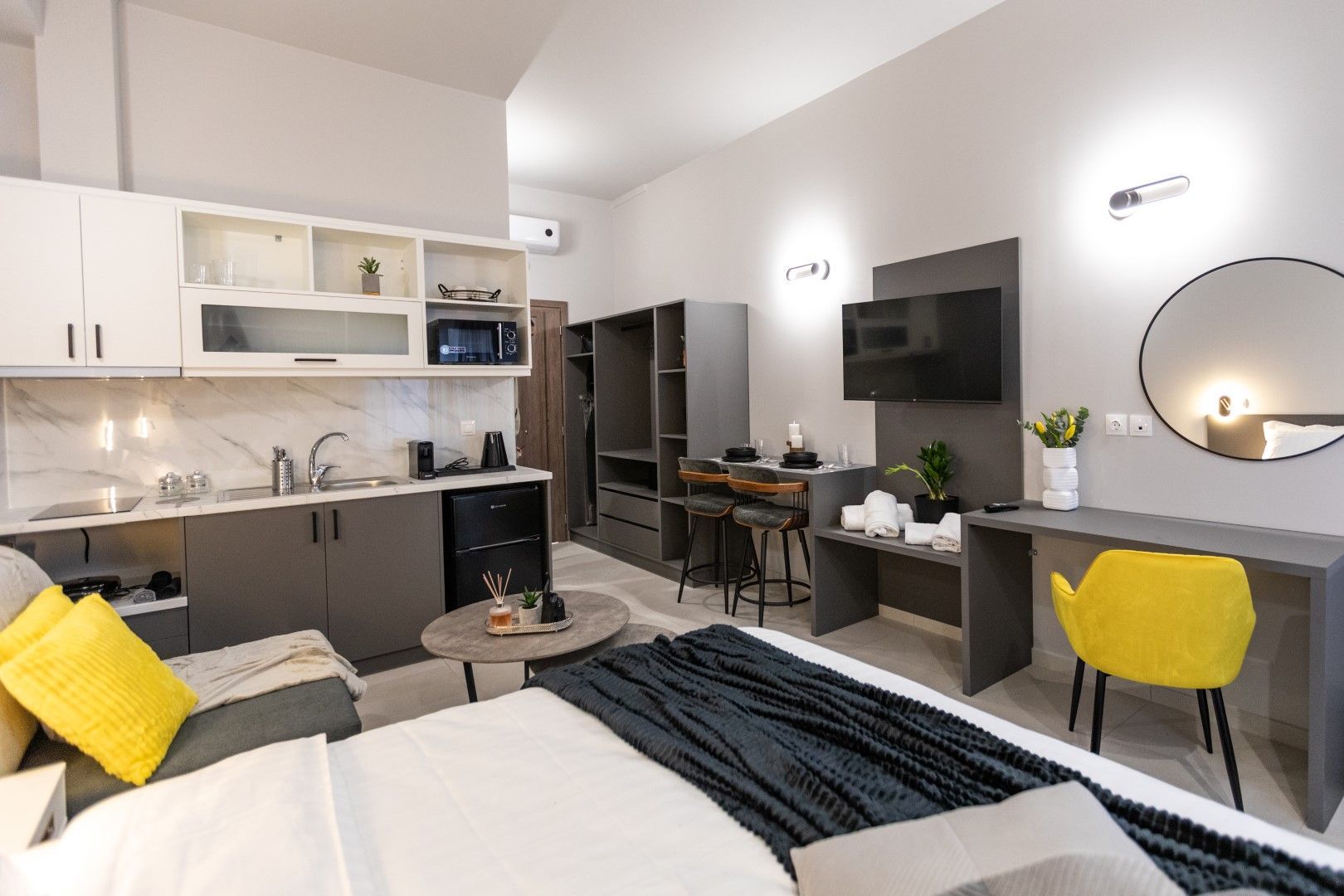 Studio apartment with gray and white kitchen, bed, desk, and round mirror.