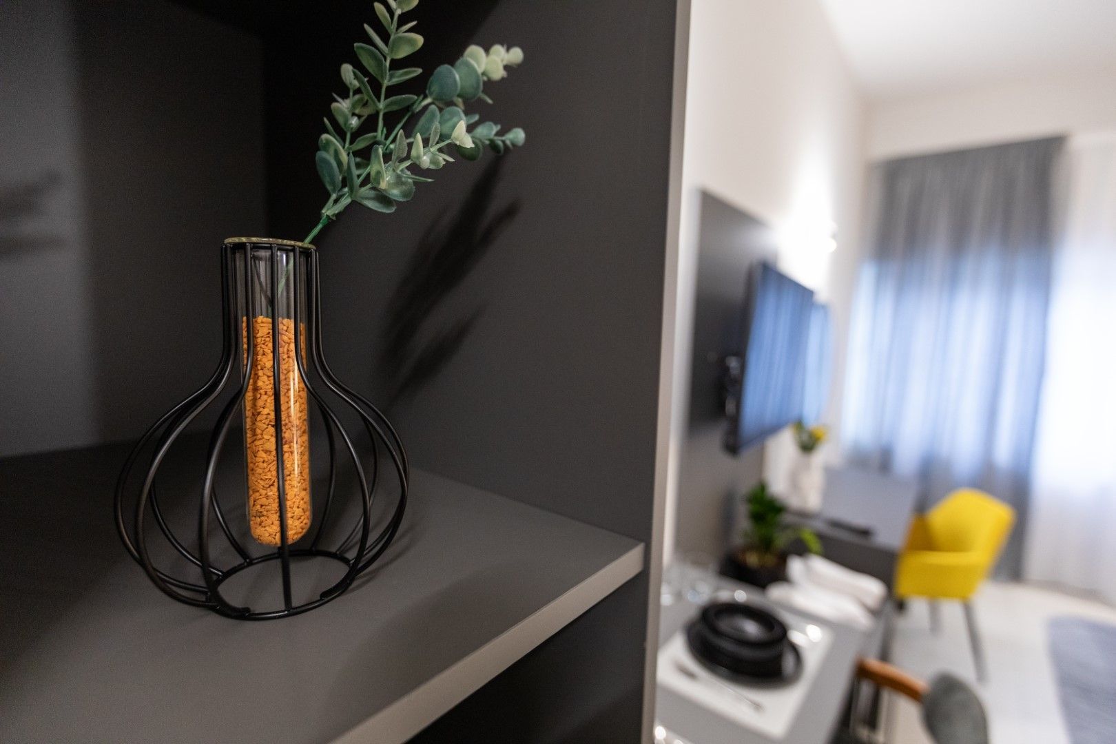 Decorative vase with sprigs on a shelf; blurry view of a room with TV and yellow chair.