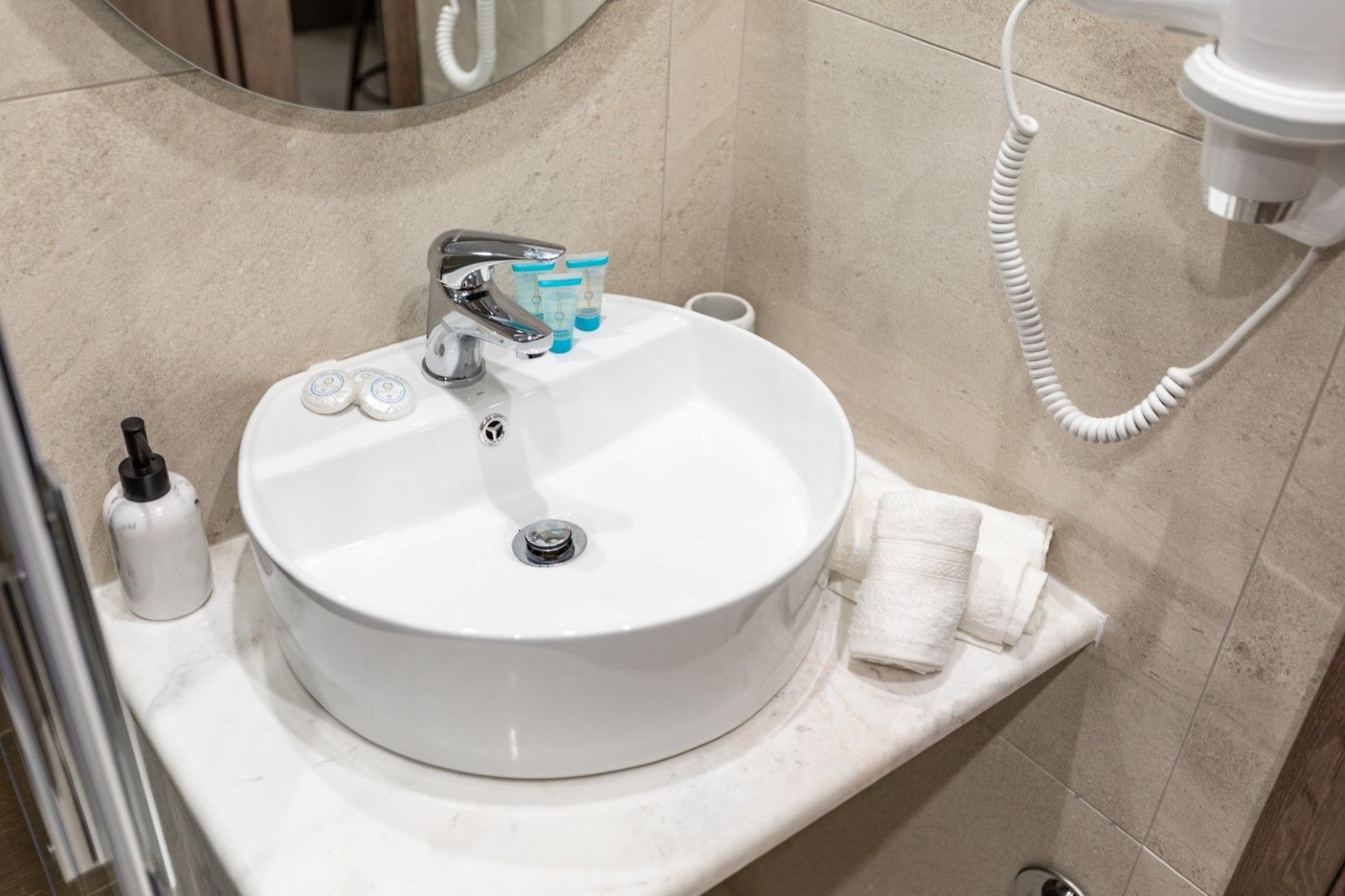 White, round bathroom sink with running water, towels, and toiletries on a marble counter. Hair dryer on the wall.