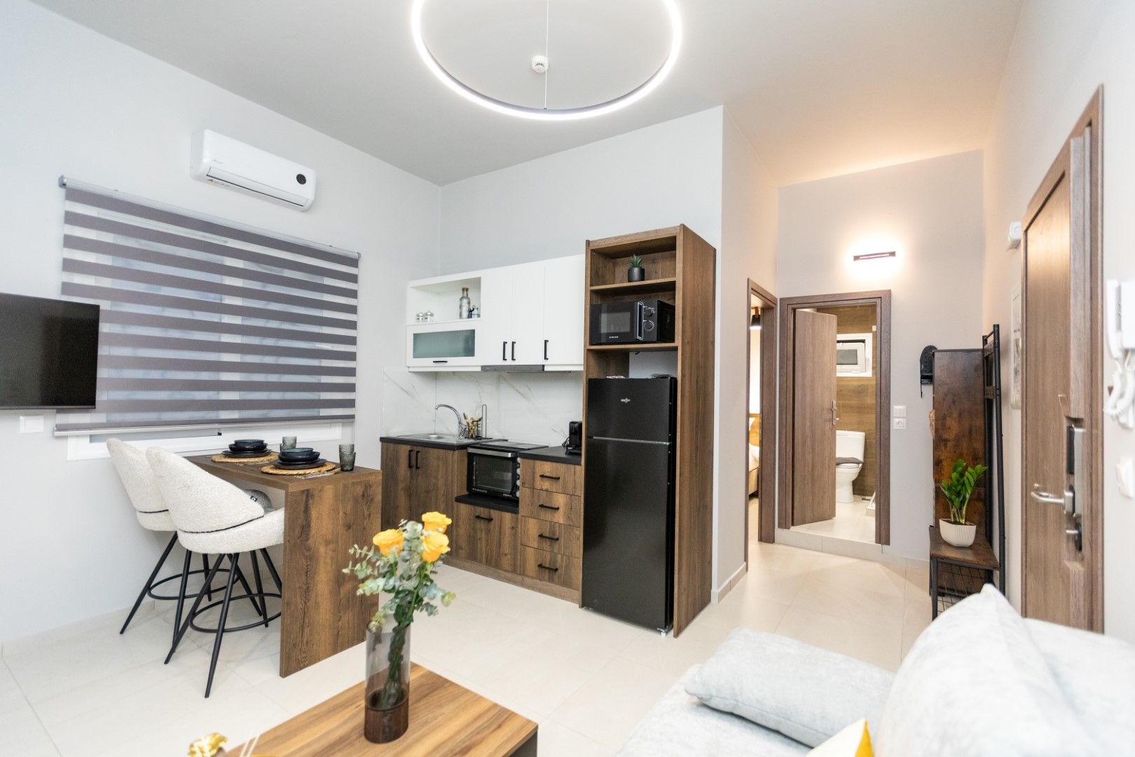 Interior of a modern apartment with kitchen, living area, and a hallway to a bathroom; light tones and wooden accents.