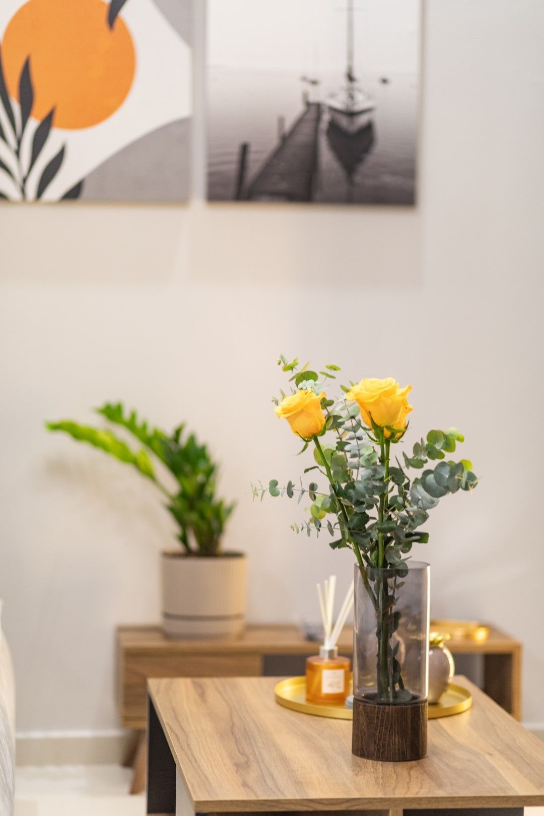 Yellow roses in a vase on a coffee table, with art and potted plant in the background.