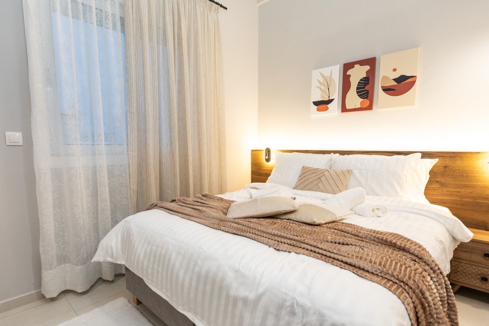 Bedroom with a bed, artwork, and window with sheer curtains. Warm, neutral tones.
