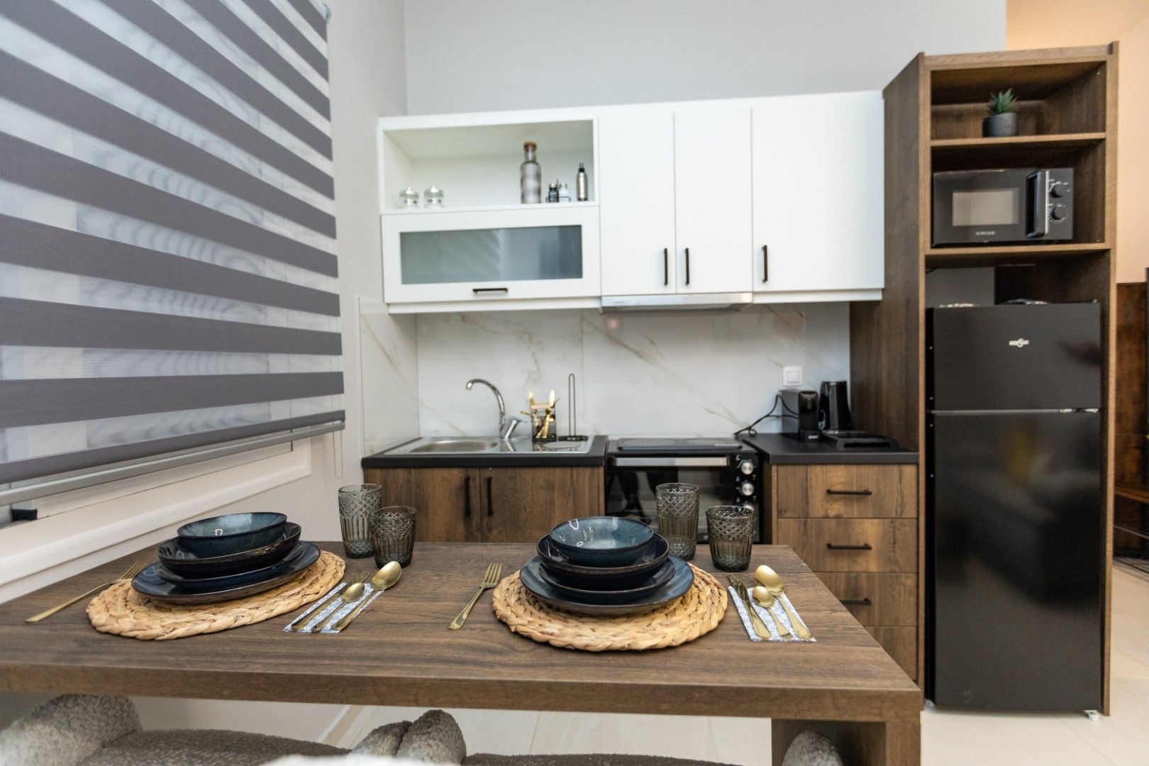 Kitchen with dining table set for two. Includes cabinets, appliances, and a window with blinds.
