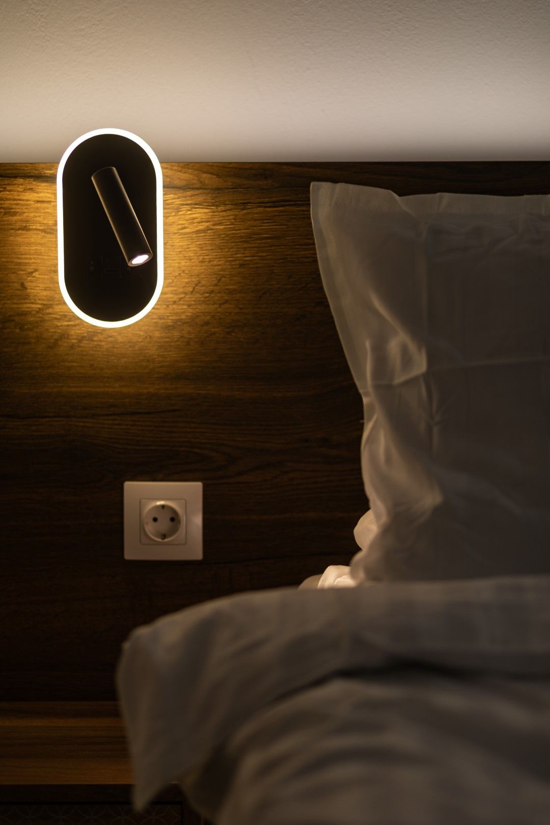 Bedside wall lamp casting light on a wooden headboard, with a pillow and power outlet.