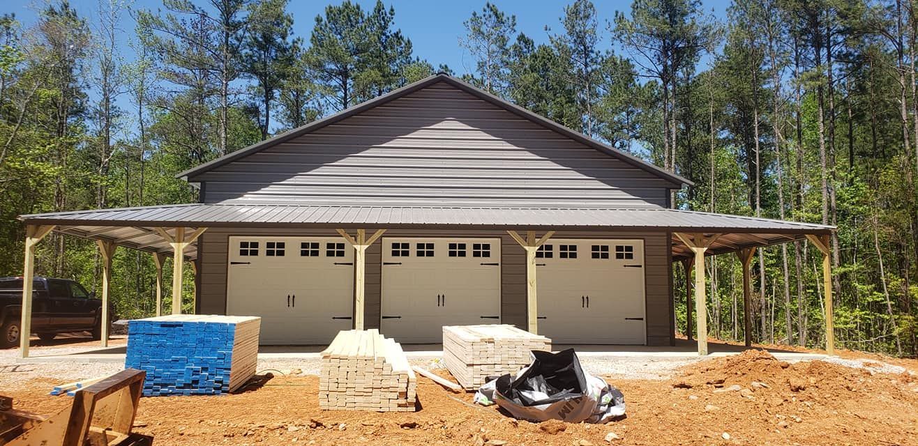A new gray three-car garage under construction, with a carport and wooded background.