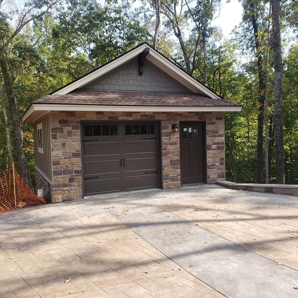 Stone-faced garage with brown doors, matching roof, and small access door; set in a wooded area with a concrete driveway.