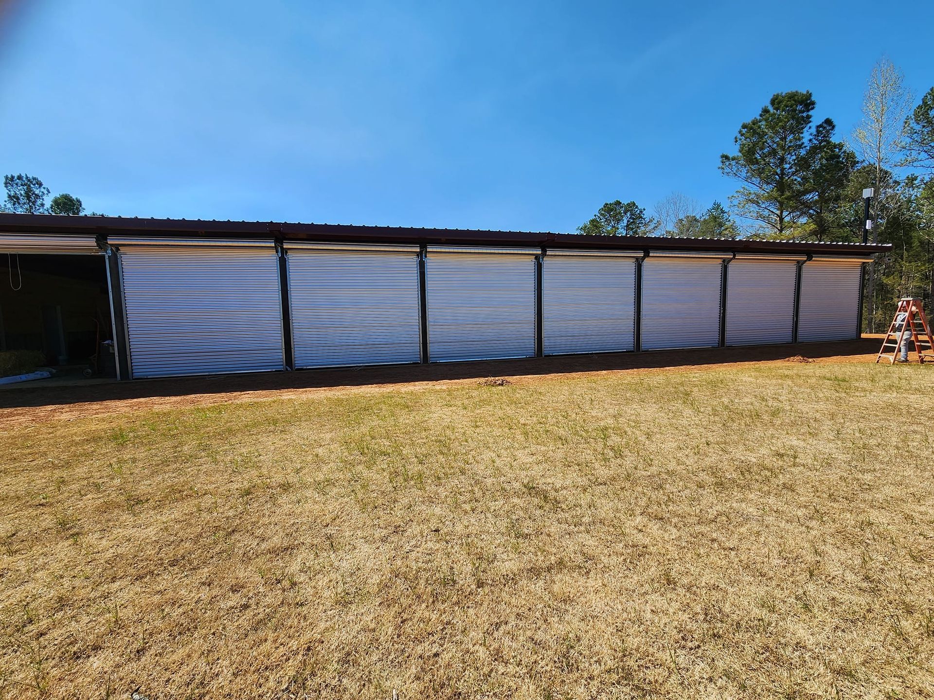 Long, open-sided shed with white, mesh walls on a sunny, grassy field. Brown roof, blue sky.