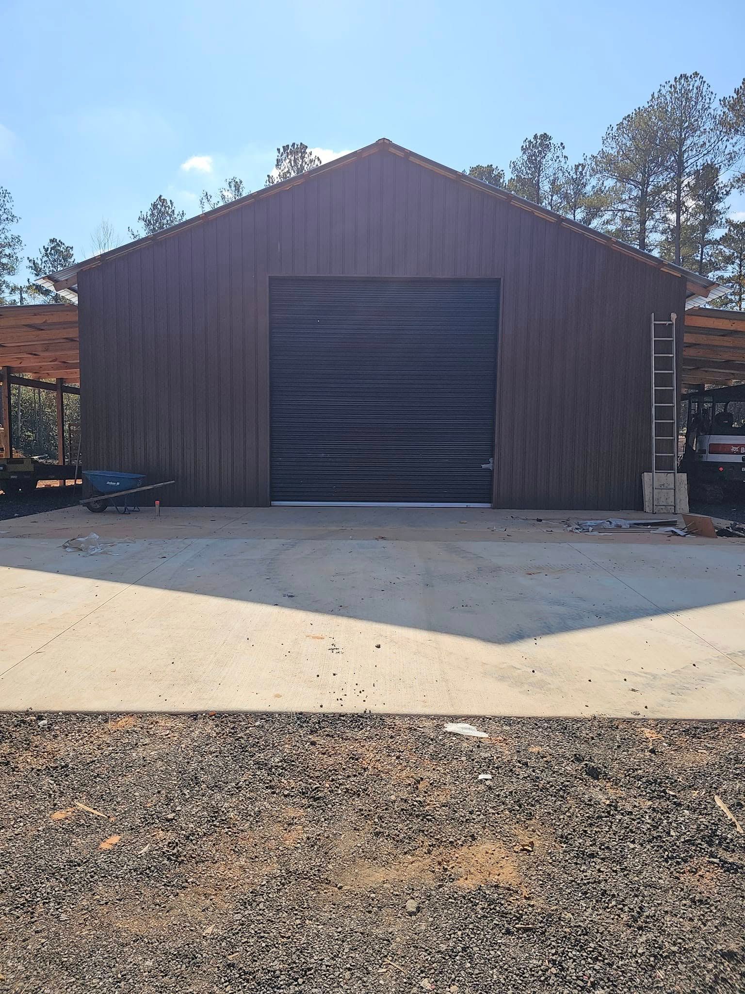 Brown barn with large black garage door, set against a blue sky, gravel lot in front.