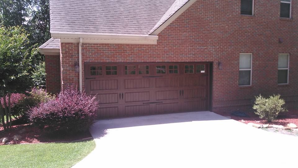 Brown garage door on a brick house, concrete driveway, and landscaping with bushes and grass.