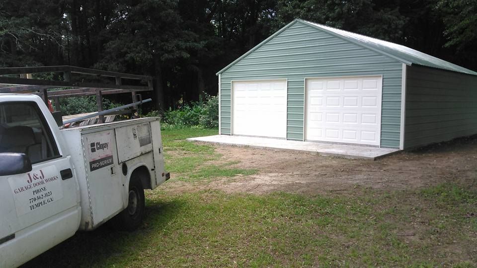 White work truck parked near a two-door green metal garage on a concrete slab, surrounded by grass and trees.