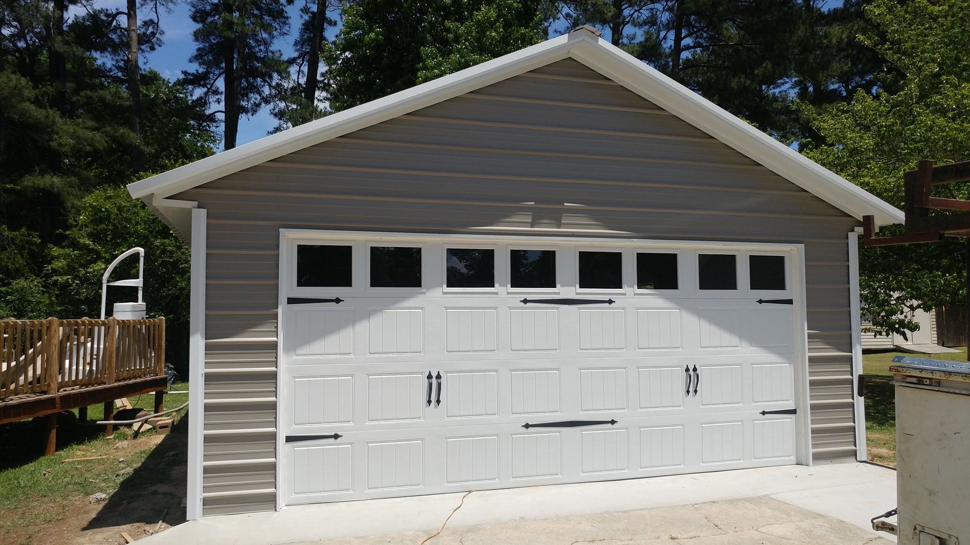 Gray and white garage with a white garage door, set in a sunny, wooded area.