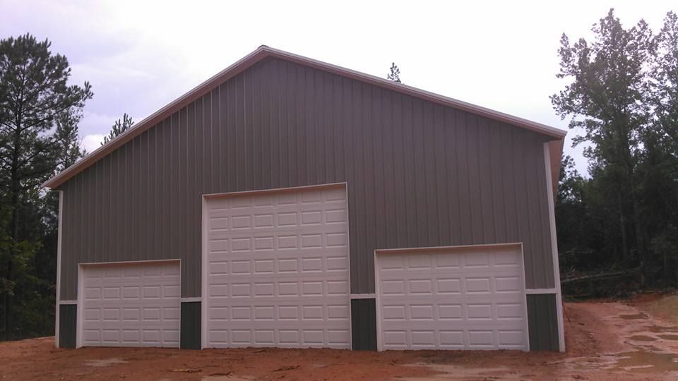 Gray metal barn with three white garage doors on a dirt path in a wooded area.