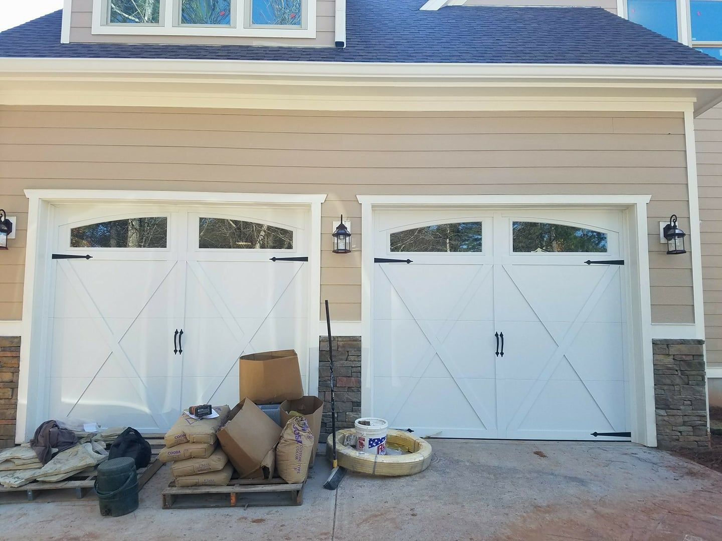 Two white garage doors with arched windows and black hardware on a tan house with stone accents.