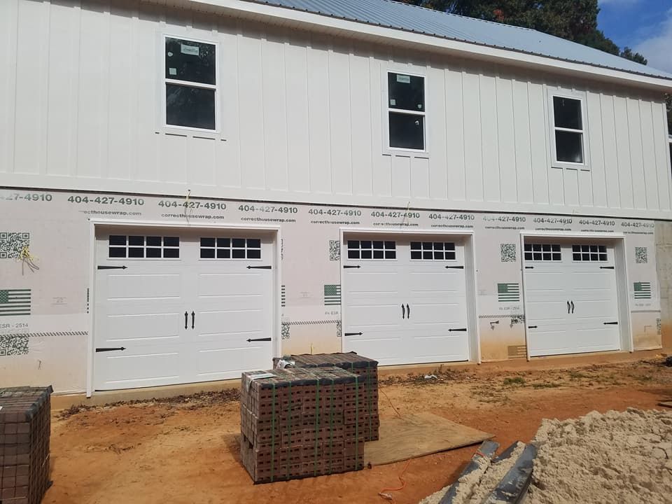 White building under construction with three garage doors. Two upper windows. Brick pile in front.