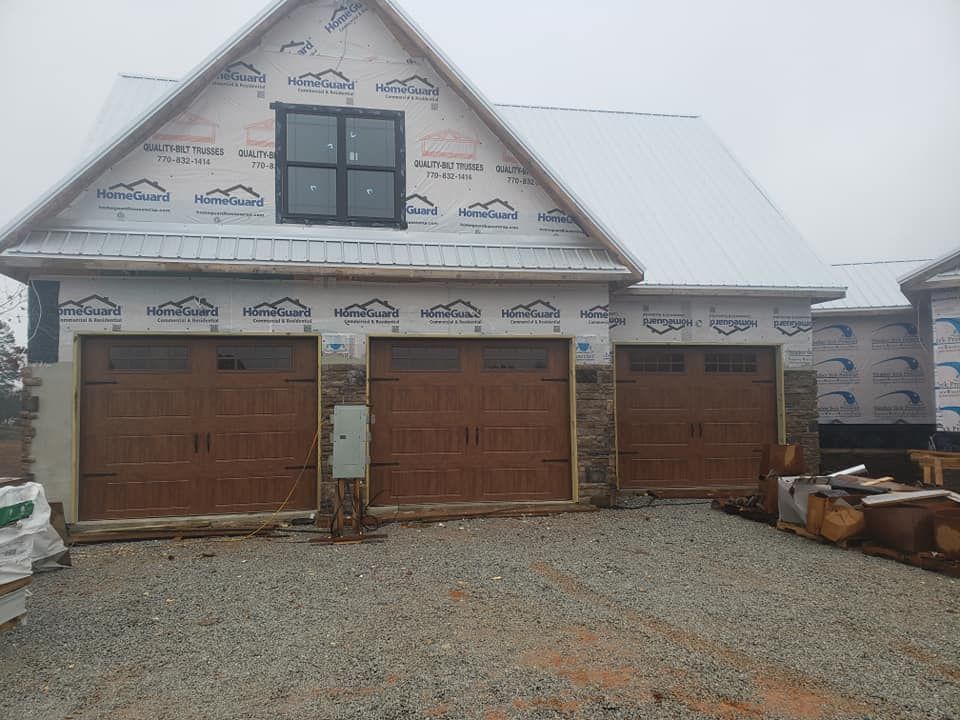 Three-car garage under construction with brown doors, stone accents, and a window. Snowy roof, gravel driveway.
